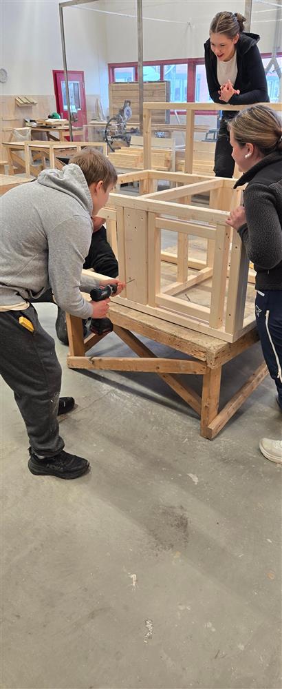 En liten gruppe elever samarbeider om å montere en trekonstruksjon på et arbeidsstativ. Én holder rammen stødig mens en annen bruker drill for å feste delene. Det er tydelig lagarbeid, og bak dem sees andre treprosjekter og verktøy i verkstedet. - Klikk for stort bilde