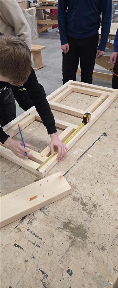 En elev sitter bøyd over en ramme på arbeidsbordet og måler opp en planke med tommestokk og blyant. Rundt ham ligger ulike verktøy, blant annet en drill og flere treplanker som skal festes. Verkstedmiljøet er aktivt og fullt av pågående prosjekter. - Klikk for stort bilde