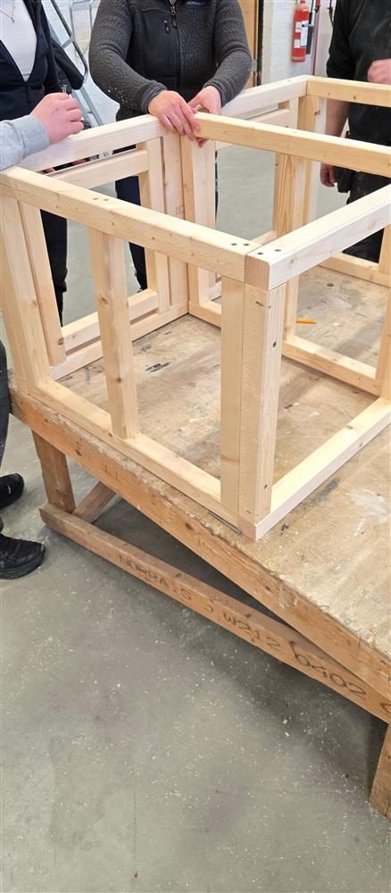En nesten ferdig trekasse eller ramme står på et stort arbeidsbord. Flere elever står rundt og holder i rammen mens de justerer og kontrollerer at alt står riktig. Konstruksjonen består av rette, lyse treplanker og ser stabil ut. - Klikk for stort bilde