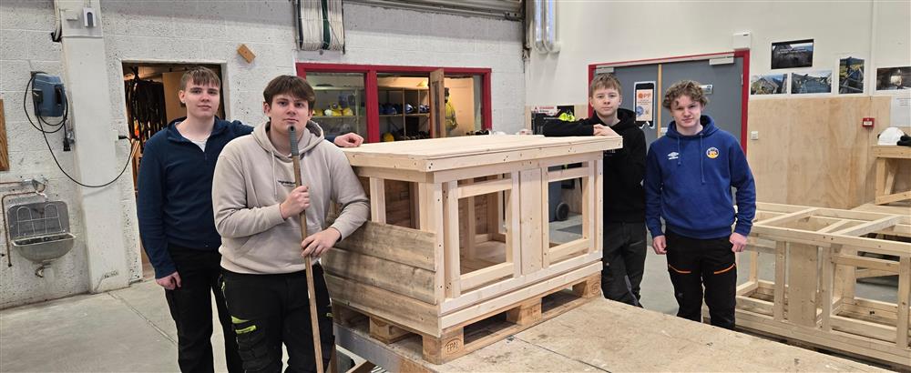 Fire elever står samlet rundt en ferdigstilt trekonstruksjon med bordplate og solide sider. Konstruksjonen står på en palle og ser ut som et større møbel eller et prosjekt som krever presist arbeid. Elevene viser frem prosjektet som et ferdig resultat etter samarbeidet. - Klikk for stort bilde