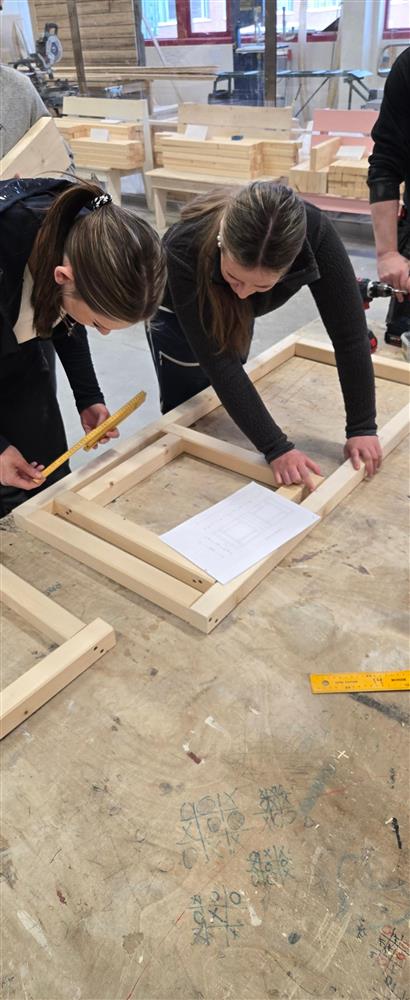 To elever jobber sammen med en del av en trekonstruksjon som ligger på bordet. Driller og skruer ligger klare rundt dem. I bakgrunnen sees større trearbeidsprosjekter, inkludert en liten bu-lignende bygning, som viser bredde i verkstedets aktiviteter. - Klikk for stort bilde